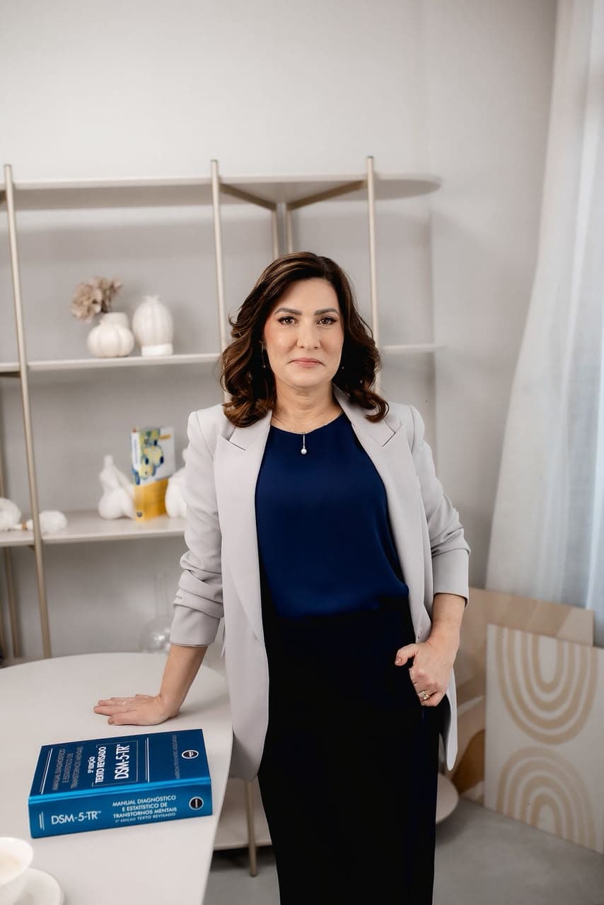 Luciana Estruque em pé, com blazer claro e o livro DSM-5 em seu ambiente profissional